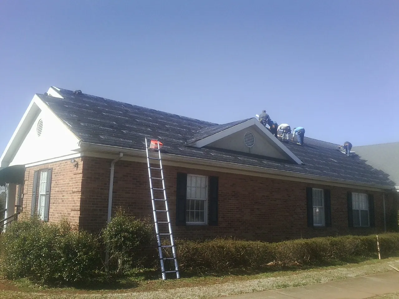 Crew tearing shingles off a brick ranch roof with a ladder set — Emergency Roof Repair work by our crew in Lavina