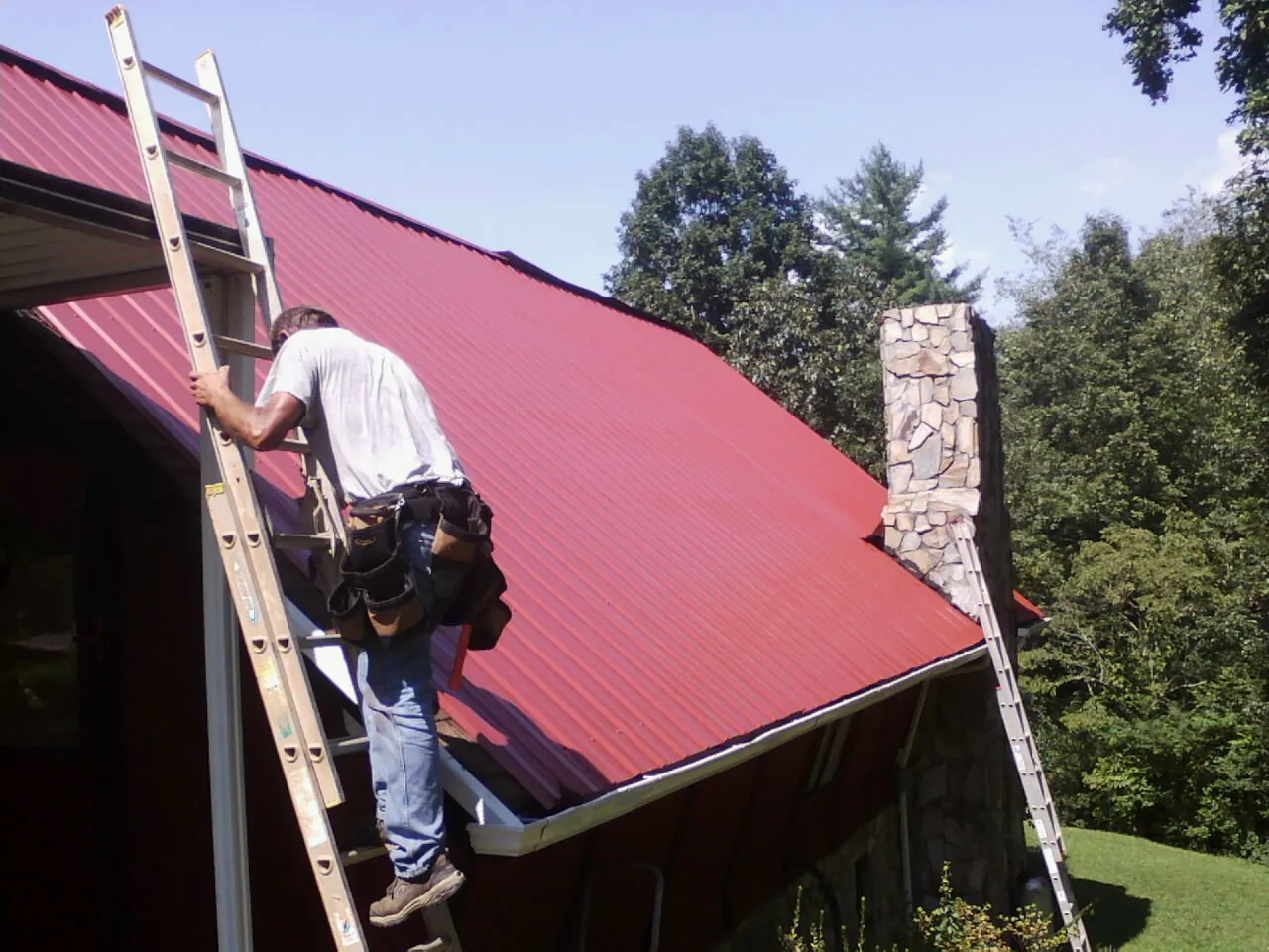 Roofer on a ladder beside a red metal roof and stone chimney — Soffit & Fascia Repair by Roofing Brothers in Lavina
